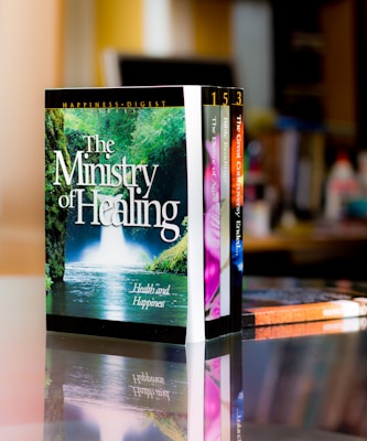 A set of colorful books is neatly arranged on a reflective surface, with the most prominent book titled 'The Ministry of Healing'. The background features a blurred indoor setting, likely a room or office.