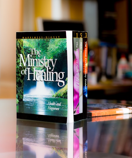 A set of colorful books is neatly arranged on a reflective surface, with the most prominent book titled 'The Ministry of Healing'. The background features a blurred indoor setting, likely a room or office.