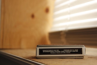 A box of playing cards labeled 'Manufactured in USA for INTERNATIONAL PLAYING CARD COMPANY LIMITED' resting on a wooden surface. Behind the box, there is a wall with light-colored wood paneling, and to the side, there are window blinds partially open, allowing light to filter through.