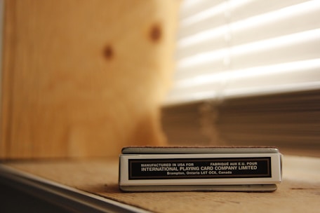 A box of playing cards labeled 'Manufactured in USA for INTERNATIONAL PLAYING CARD COMPANY LIMITED' resting on a wooden surface. Behind the box, there is a wall with light-colored wood paneling, and to the side, there are window blinds partially open, allowing light to filter through.