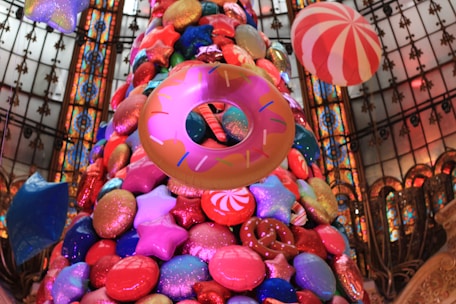 A large, colorful Christmas tree adorned with oversized, whimsical decorations including stars, donuts, and candy-like ornaments. The background features a decorative stained glass ceiling with intricate patterns.