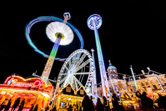 A vibrant nighttime carnival scene with brightly lit rides and attractions. A Ferris wheel dominates the center, surrounded by various thrill rides with colorful light trails, including a spinning swing ride creating circular light patterns. In the foreground, there is a concession stand that seems to specialize in sweets. People are seen as silhouettes, enjoying the festive atmosphere under a clear night sky.
