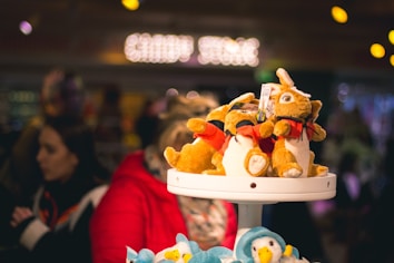 Several plush kangaroo toys are displayed on a white rack in a store. The background is blurred, showing people and a sign that seems to read 'Candy Store' with bright lights.