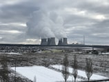 A large industrial complex with multiple cooling towers emitting steam into an overcast sky. In the foreground, a snowy field and a small town with scattered buildings are visible.