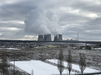 A large industrial complex with multiple cooling towers emitting steam into an overcast sky. In the foreground, a snowy field and a small town with scattered buildings are visible.