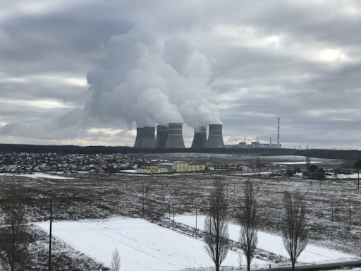 A large industrial complex with multiple cooling towers emitting steam into an overcast sky. In the foreground, a snowy field and a small town with scattered buildings are visible.