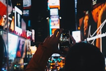 A bustling urban scene with bright, colorful billboards and neon lights illuminating the night. A person is capturing the vibrant environment with a smartphone, held up close to their face. The atmosphere is lively and dynamic, indicative of a busy, popular location.