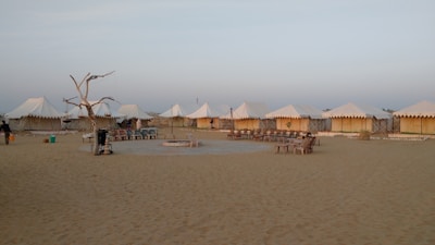 A desert campsite with several white tents arranged in a row. The area is sandy, and there is a ring of seating around a central fire pit. A dry, leafless tree stands to one side, surrounded by some basic outdoor furniture.