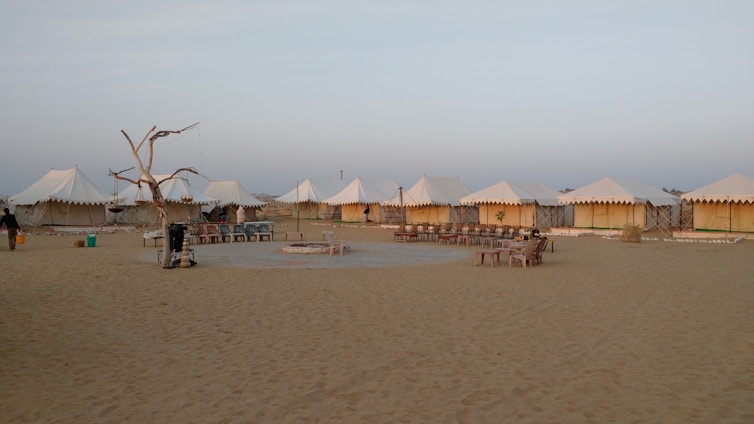 A desert campsite with several white tents arranged in a row. The area is sandy, and there is a ring of seating around a central fire pit. A dry, leafless tree stands to one side, surrounded by some basic outdoor furniture.
