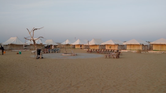 A desert campsite with several white tents arranged in a row. The area is sandy, and there is a ring of seating around a central fire pit. A dry, leafless tree stands to one side, surrounded by some basic outdoor furniture.