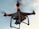 A drone with a camera attached is flying against a backdrop of a clear sky. The drone has red lights indicating its active status, and its rotors are visible on each corner, gently blurred to show motion.
