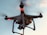 A drone with a camera attached is flying against a backdrop of a clear sky. The drone has red lights indicating its active status, and its rotors are visible on each corner, gently blurred to show motion.