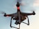 A drone with a camera attached is flying against a backdrop of a clear sky. The drone has red lights indicating its active status, and its rotors are visible on each corner, gently blurred to show motion.