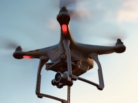 A drone with a camera attached is flying against a backdrop of a clear sky. The drone has red lights indicating its active status, and its rotors are visible on each corner, gently blurred to show motion.