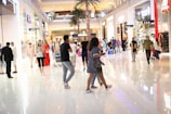 A busy shopping mall with people walking, talking, and using their phones. The environment is bright with reflective floors, and stores line the sides of the space. A large palm tree decorates the area, and the ambiance suggests a lively and bustling atmosphere.