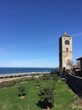 A stone clock tower stands prominently against a clear blue sky, surrounded by a well-maintained green lawn with neatly trimmed bushes and small trees. In the background, an expansive view of the ocean is visible with a stretch of highway running parallel to the coastline.