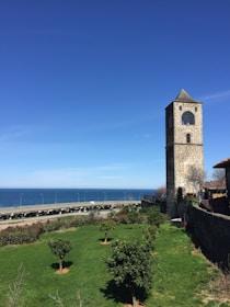 A stone clock tower stands prominently against a clear blue sky, surrounded by a well-maintained green lawn with neatly trimmed bushes and small trees. In the background, an expansive view of the ocean is visible with a stretch of highway running parallel to the coastline.