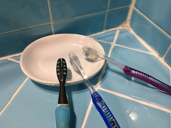 A white dish is resting on a tiled blue surface, with three toothbrushes placed on it. The toothbrushes have different colored handles: black, blue, and purple. The surrounding area features blue tiles with visible grout lines.