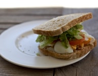 A sandwich sits on a white plate placed on a wooden surface. It includes layers of lettuce, sliced tomatoes, boiled egg, and cheese, all between two slices of whole wheat bread.