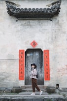 A woman stands in front of an old, rustic wooden door adorned with red banners featuring Chinese calligraphy. She wears a hat, sweater, and slacks, and carries a backpack, posing against the backdrop of an aged, weathered wall with classic architectural detailing.