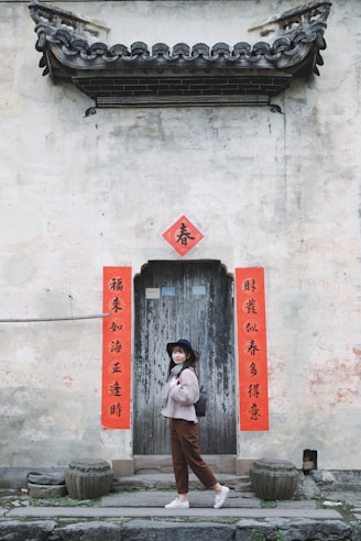 A woman stands in front of an old, rustic wooden door adorned with red banners featuring Chinese calligraphy. She wears a hat, sweater, and slacks, and carries a backpack, posing against the backdrop of an aged, weathered wall with classic architectural detailing.