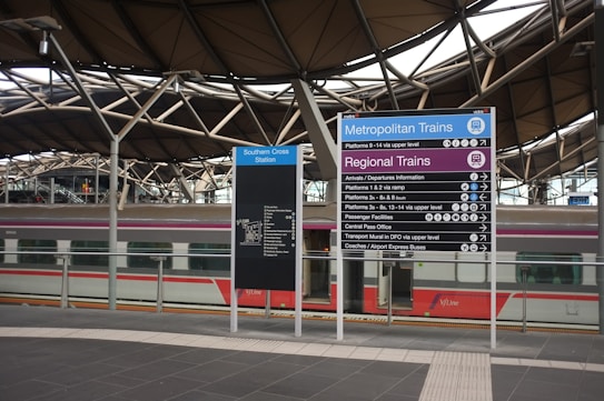 A modern train station with an intricate roof structure. Prominent signage directs passengers to metropolitan and regional train platforms. A train with a sleek design is visible underneath the roof, and platforms are clearly outlined.