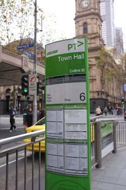 A public transportation information board is prominently displayed in an urban setting. The board is predominantly green and provides details for tram services at the Town Hall stop on Collins Street. Behind the board is a historic clock tower building and a busy street. People can be seen walking along the sidewalk, and there is a yellow taxi nearby.