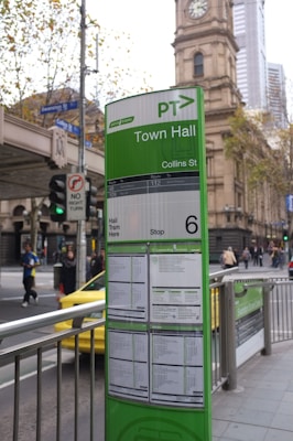 A public transportation information board is prominently displayed in an urban setting. The board is predominantly green and provides details for tram services at the Town Hall stop on Collins Street. Behind the board is a historic clock tower building and a busy street. People can be seen walking along the sidewalk, and there is a yellow taxi nearby.