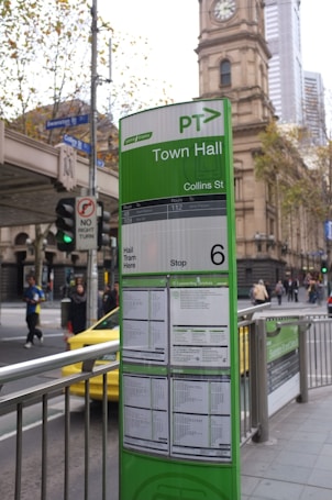A public transportation information board is prominently displayed in an urban setting. The board is predominantly green and provides details for tram services at the Town Hall stop on Collins Street. Behind the board is a historic clock tower building and a busy street. People can be seen walking along the sidewalk, and there is a yellow taxi nearby.