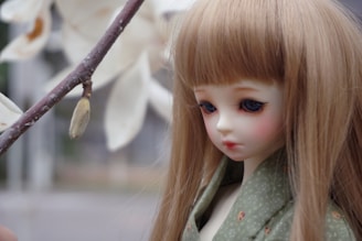 A doll with long, straight, blonde hair and detailed facial features is positioned next to a branch with a flower bud. The doll is dressed in a green garment with subtle patterns.