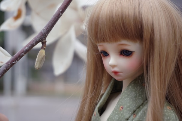 A doll with long, straight, blonde hair and detailed facial features is positioned next to a branch with a flower bud. The doll is dressed in a green garment with subtle patterns.