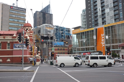 An urban cityscape featuring a diverse mix of architectural styles, including modern glass buildings and Victorian-style structures. Visible are several vans driving across a street intersection, with prominent signage for RMIT University and Hard Rock Cafe. The scene is bustling with various buildings and urban elements such as traffic lights and power lines.