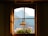 A picturesque view of mountains is visible through a window with a wooden frame and curtains. A hanging lamp is centered in the foreground, and blooming flowers in a planter adorn the windowsill, adding a touch of nature to the cozy interior ambiance.