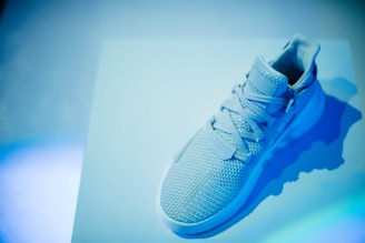 A single sneaker with a woven texture and laces is placed on a surface, surrounded by a blue-tinted light. The sneaker appears to have a modern and sporty design.
