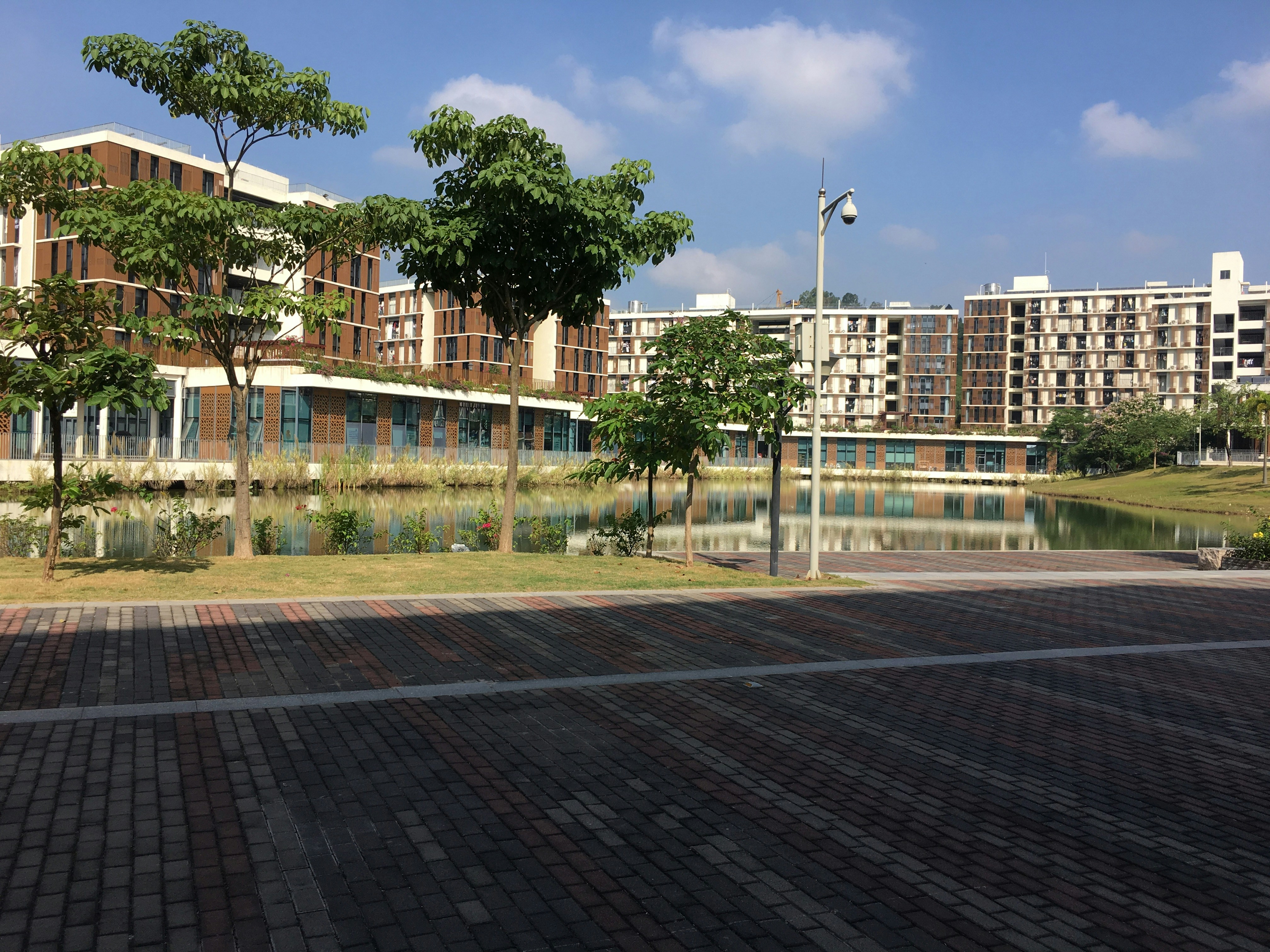 A modern residential complex is seen with multiple buildings featuring a combination of brown and beige tones. There is a landscaped area with several green trees and a calm water body reflecting the buildings. The foreground includes a paved pathway with a mix of brown and grey bricks. The sky is clear with a few scattered clouds, indicating a sunny day.