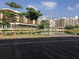 A modern residential complex is seen with multiple buildings featuring a combination of brown and beige tones. There is a landscaped area with several green trees and a calm water body reflecting the buildings. The foreground includes a paved pathway with a mix of brown and grey bricks. The sky is clear with a few scattered clouds, indicating a sunny day.