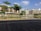 A modern residential complex is seen with multiple buildings featuring a combination of brown and beige tones. There is a landscaped area with several green trees and a calm water body reflecting the buildings. The foreground includes a paved pathway with a mix of brown and grey bricks. The sky is clear with a few scattered clouds, indicating a sunny day.