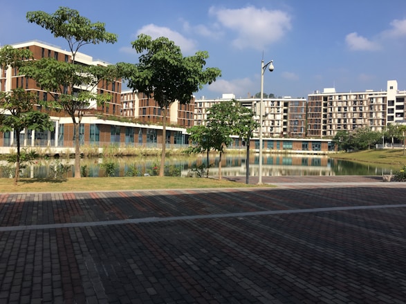 A modern residential complex is seen with multiple buildings featuring a combination of brown and beige tones. There is a landscaped area with several green trees and a calm water body reflecting the buildings. The foreground includes a paved pathway with a mix of brown and grey bricks. The sky is clear with a few scattered clouds, indicating a sunny day.