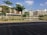 A modern residential complex is seen with multiple buildings featuring a combination of brown and beige tones. There is a landscaped area with several green trees and a calm water body reflecting the buildings. The foreground includes a paved pathway with a mix of brown and grey bricks. The sky is clear with a few scattered clouds, indicating a sunny day.