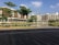 A modern residential complex is seen with multiple buildings featuring a combination of brown and beige tones. There is a landscaped area with several green trees and a calm water body reflecting the buildings. The foreground includes a paved pathway with a mix of brown and grey bricks. The sky is clear with a few scattered clouds, indicating a sunny day.