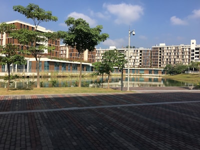 A modern residential complex is seen with multiple buildings featuring a combination of brown and beige tones. There is a landscaped area with several green trees and a calm water body reflecting the buildings. The foreground includes a paved pathway with a mix of brown and grey bricks. The sky is clear with a few scattered clouds, indicating a sunny day.