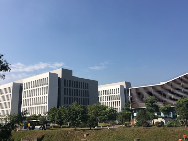 A cluster of modern office buildings with large windows and a clean, geometric design. The buildings are surrounded by well-maintained greenery and trees. A clear blue sky is visible above. A bus is parked near one of the buildings.