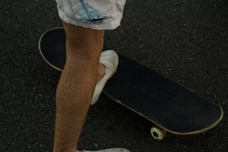 A close-up view of a person's legs wearing white shorts standing next to a skateboard on a textured surface. The skateboard is positioned diagonally, with one foot partially resting on its deck.