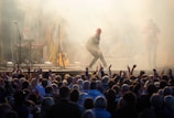 A lively concert scene with a performer in a red hat energetically engaging with a large, enthusiastic audience. The stage is surrounded by smoke effects, with musicians playing instruments like a keyboard and guitar in the background. The crowd is visible in the foreground, raising their hands in excitement.
