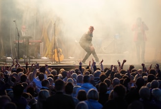 A lively concert scene with a performer in a red hat energetically engaging with a large, enthusiastic audience. The stage is surrounded by smoke effects, with musicians playing instruments like a keyboard and guitar in the background. The crowd is visible in the foreground, raising their hands in excitement.