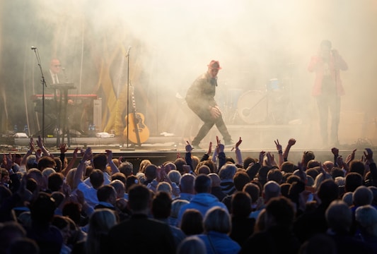 A lively concert scene with a performer in a red hat energetically engaging with a large, enthusiastic audience. The stage is surrounded by smoke effects, with musicians playing instruments like a keyboard and guitar in the background. The crowd is visible in the foreground, raising their hands in excitement.