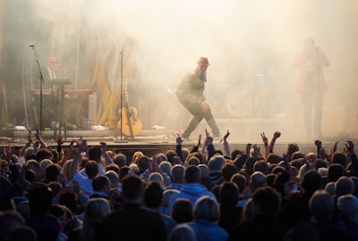 A lively concert scene with a performer in a red hat energetically engaging with a large, enthusiastic audience. The stage is surrounded by smoke effects, with musicians playing instruments like a keyboard and guitar in the background. The crowd is visible in the foreground, raising their hands in excitement.