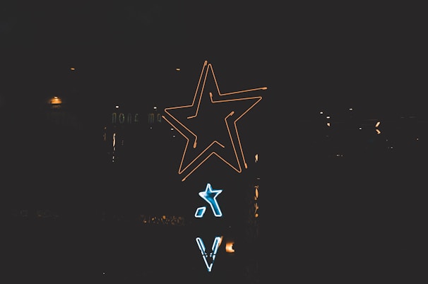 Neon signs shaped like stars and letters are illuminated against a dark background. The larger star glows in yellow, while a smaller one shines in blue beneath it.