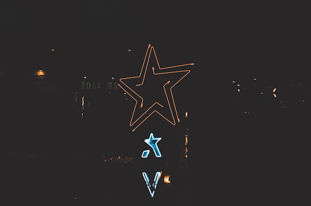 Neon signs shaped like stars and letters are illuminated against a dark background. The larger star glows in yellow, while a smaller one shines in blue beneath it.