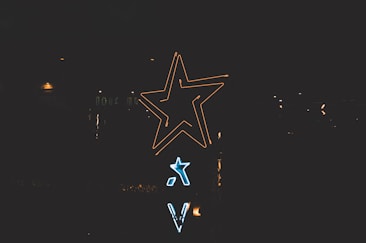 Neon signs shaped like stars and letters are illuminated against a dark background. The larger star glows in yellow, while a smaller one shines in blue beneath it.
